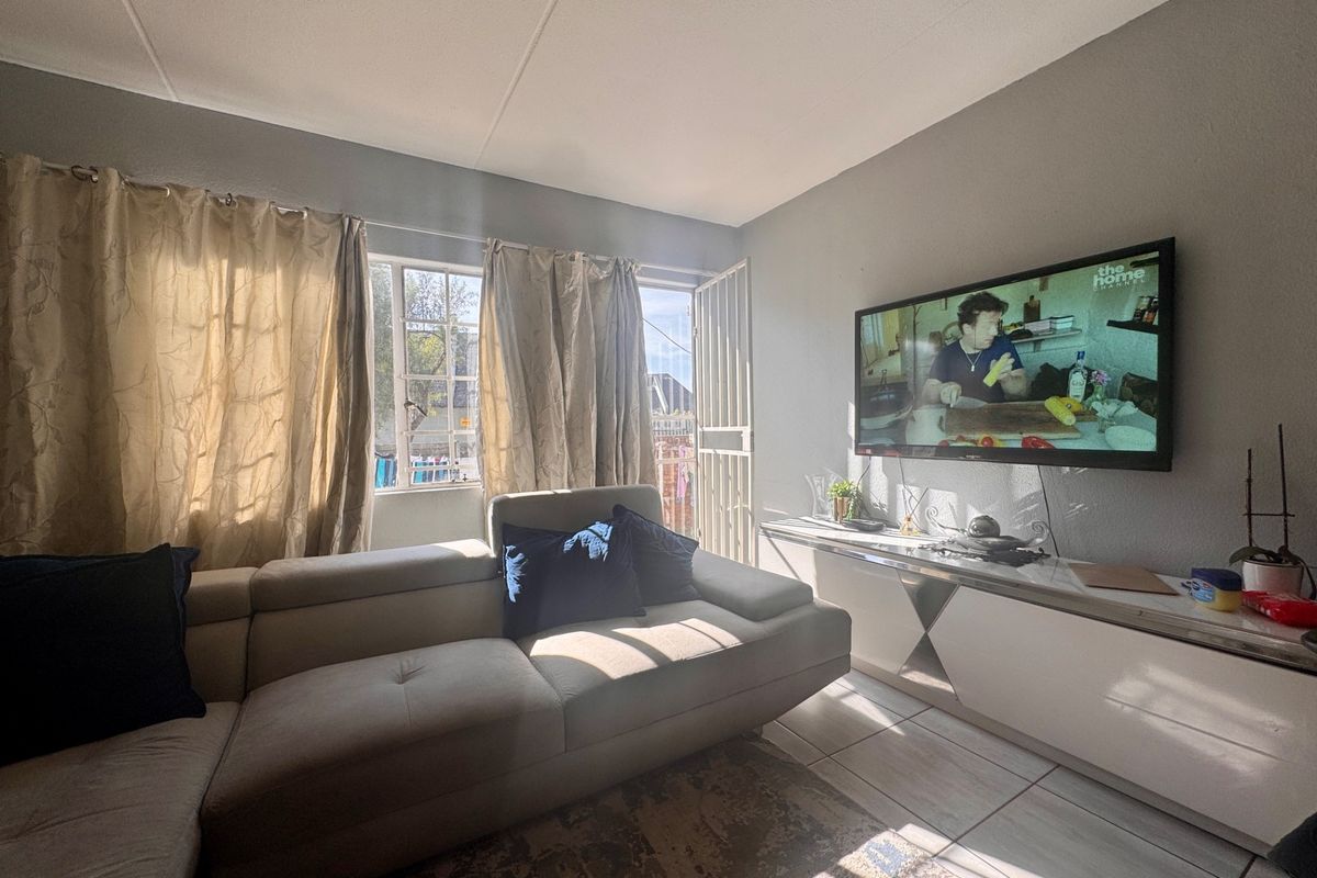 A bright living room featuring tiled flooring, large windows with natural light, and a security gate leading to the exterior.