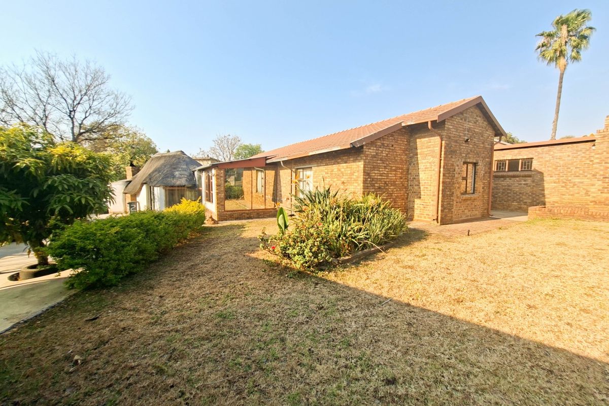 Rear view of a face brick house featuring a tiled roof, a small paved patio area, and an adjacent thatched roof structure in the garden.