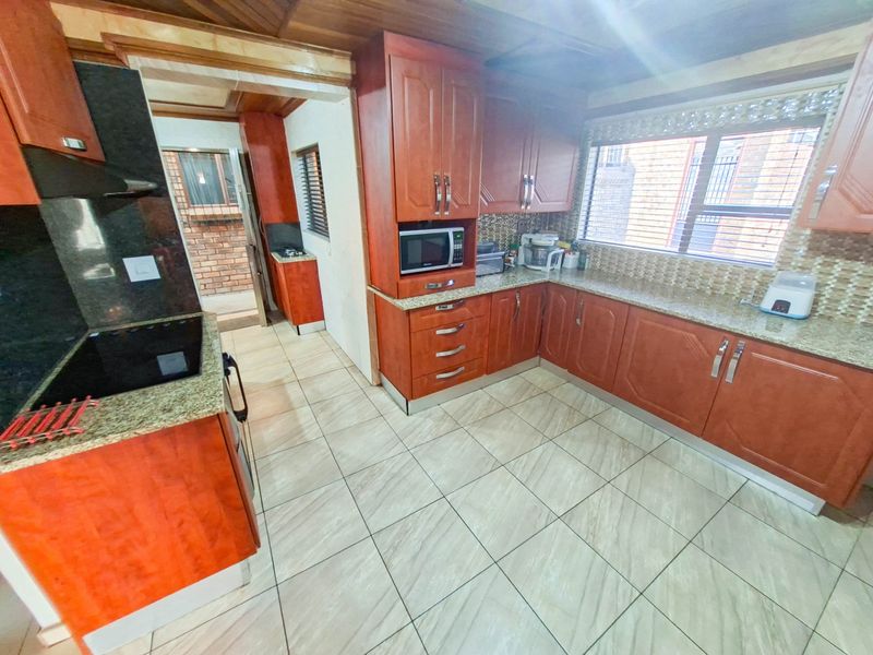 A functional kitchen featuring warm-toned wooden cabinetry, granite countertops, a mosaic tile backsplash, and tiled flooring, with an adjacent scullery area visible.
