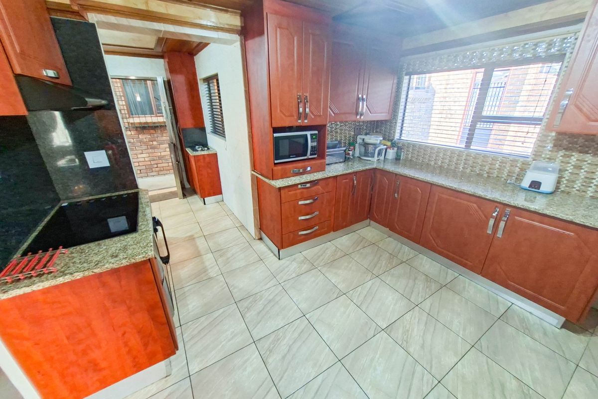 A functional kitchen featuring warm-toned wooden cabinetry, granite countertops, a mosaic tile backsplash, and tiled flooring, with an adjacent scullery area visible.