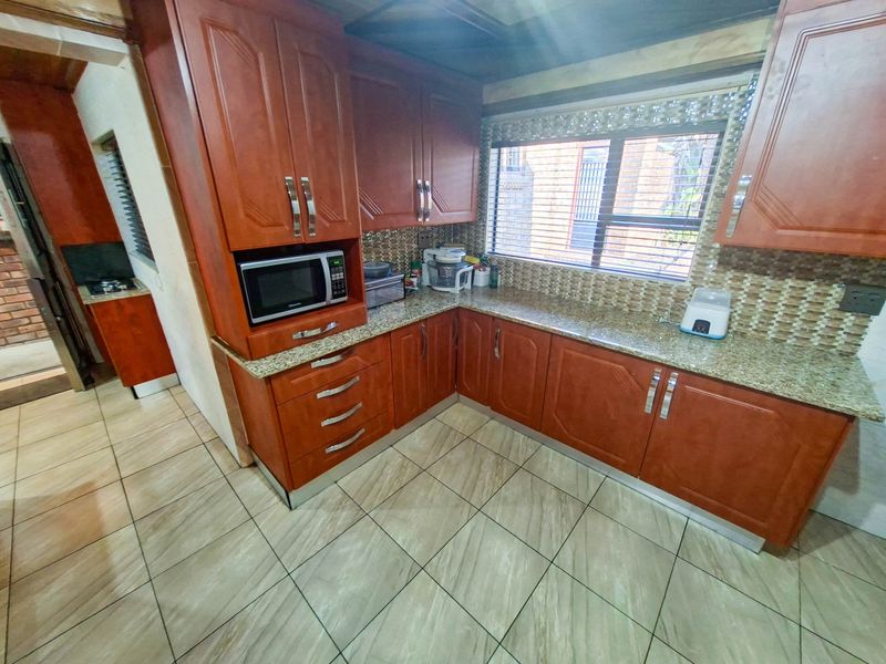 A functional kitchen featuring warm-toned wooden cabinetry, granite countertops, a mosaic tile backsplash, and tiled flooring with an adjacent scullery area.