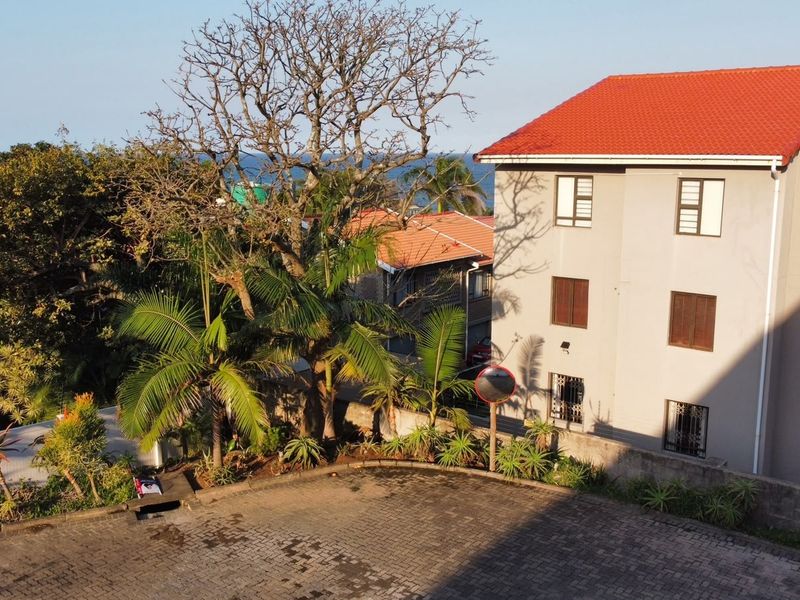 An elevated aerial view of the apartment complex exterior, featuring a paved driveway, landscaped garden area, and the building's tiled roof and facade.