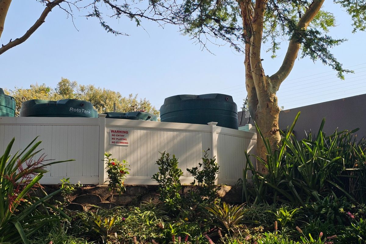 Rear garden area featuring a landscaped garden, retaining wall, and a secure vinyl fence enclosing multiple water storage tanks.