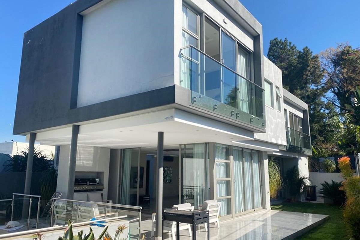 Modern double-story rear elevation featuring a spacious tiled patio, built-in braai, and glass-enclosed balconies with contemporary architectural finishes.
