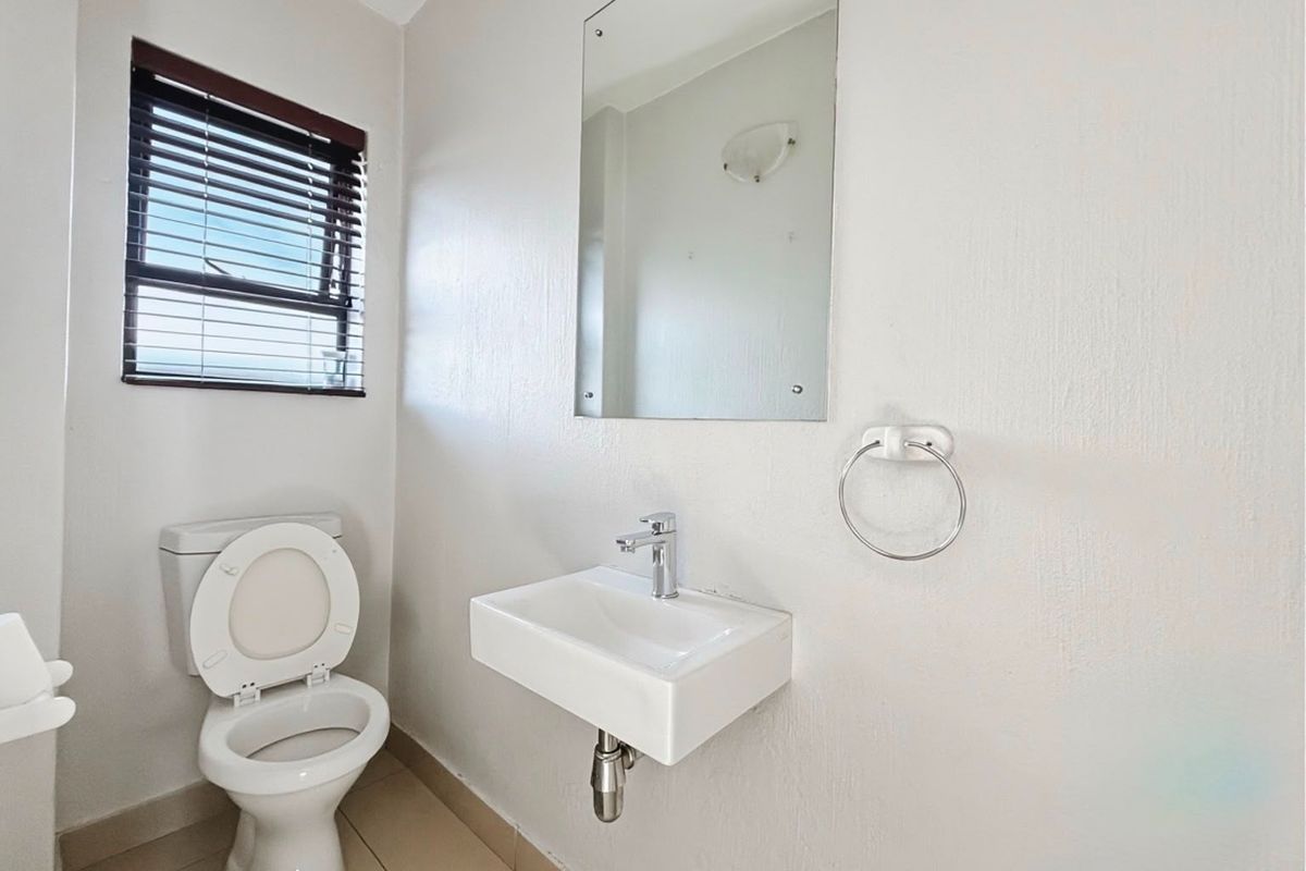 A clean and functional bathroom featuring a wall-mounted basin, chrome fixtures, and a toilet, finished with neutral wall tones and a window blind.