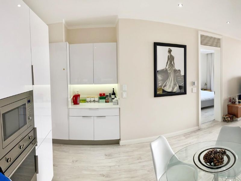 Modern open-plan living area featuring sleek white cabinetry, integrated appliances, recessed lighting, and light-toned laminate flooring, leading into an adjacent bedroom.