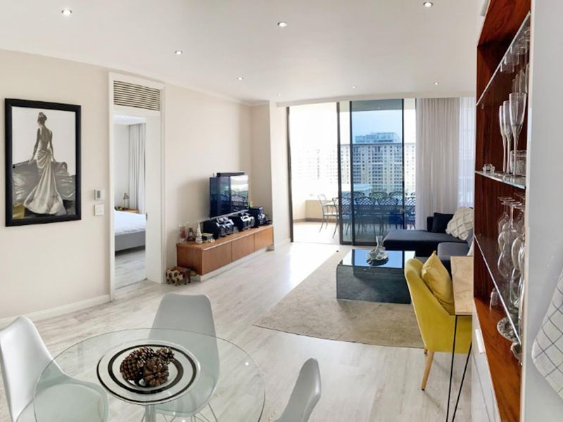 Bright open-plan living and dining area featuring modern laminate flooring, recessed lighting, and seamless access to a private balcony through large sliding glass doors.