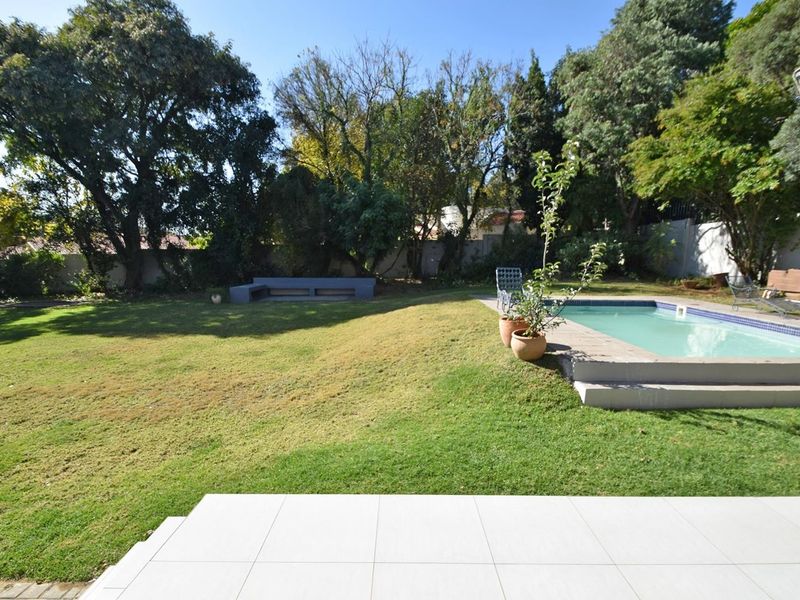 A spacious backyard featuring a swimming pool, a tiled patio area, and a built-in bench set within a landscaped garden.