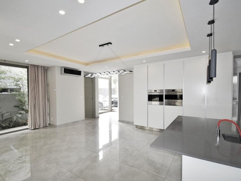 Modern open-plan living and kitchen area featuring high-gloss cabinetry, built-in double ovens, stone island countertop, recessed ceiling lighting, and large sliding doors to the garden.