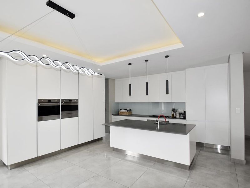 Modern kitchen featuring sleek white cabinetry, a central island with stone countertops, integrated double ovens, and contemporary pendant lighting.