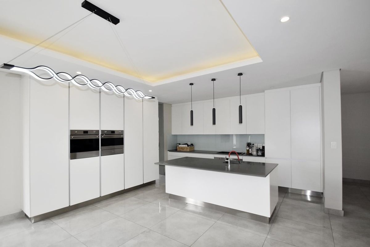 Modern kitchen featuring sleek white cabinetry, a central island with stone countertops, integrated double ovens, and contemporary pendant lighting.