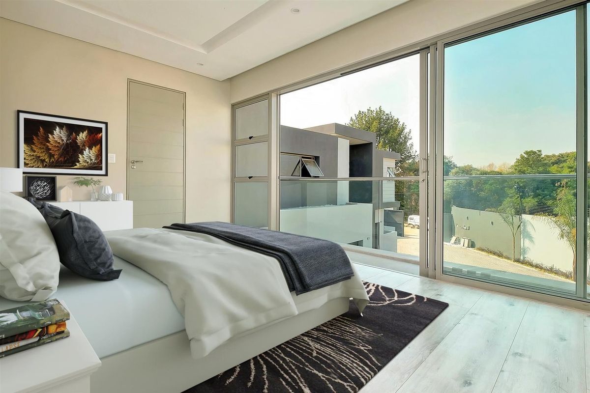 Modern bedroom featuring light laminate flooring, recessed ceiling lighting, and large sliding glass doors opening onto a balcony with a glass balustrade.