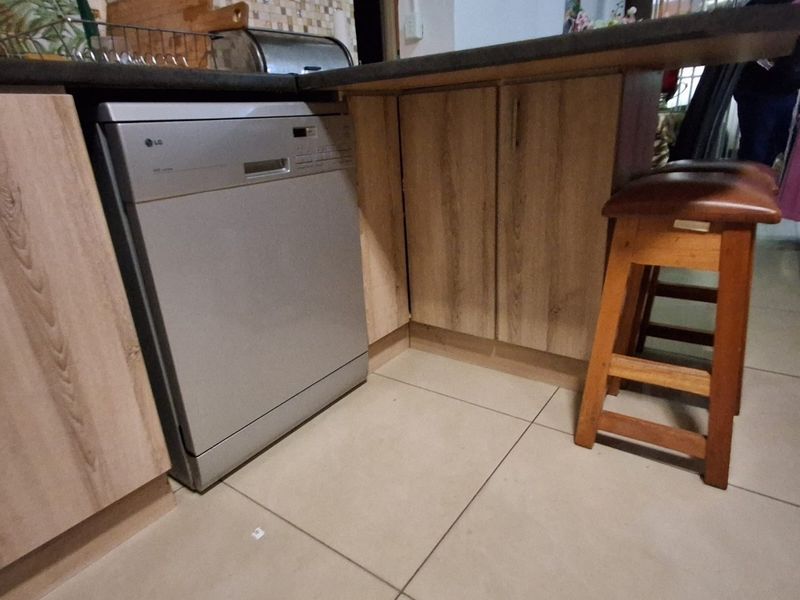 A functional kitchen area featuring wood-look cabinetry, dark countertops, a mosaic tile backsplash, and integrated dishwasher with tiled flooring.