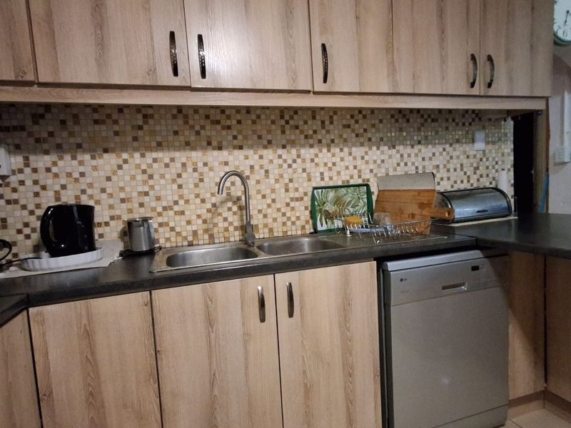 A functional kitchen featuring light wood-look cabinetry, a mosaic tile backsplash, stainless steel sink, and integrated dishwasher with neutral tiled flooring.