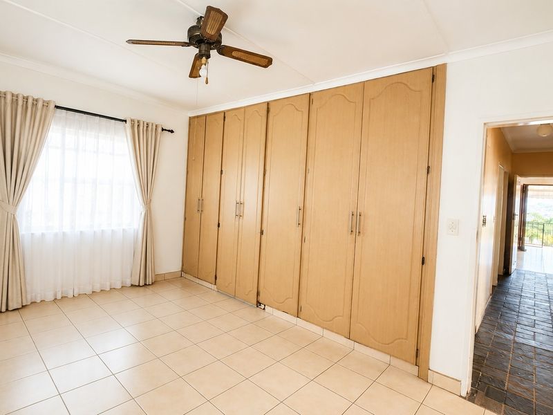 A spacious bedroom featuring neutral tiled flooring, extensive built-in wooden cupboards, and a ceiling fan, offering a clean and functional living space.