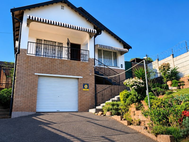 Front elevation of a two-story brick house featuring a single garage, an upper-level balcony with striped awnings, and a landscaped terraced garden.