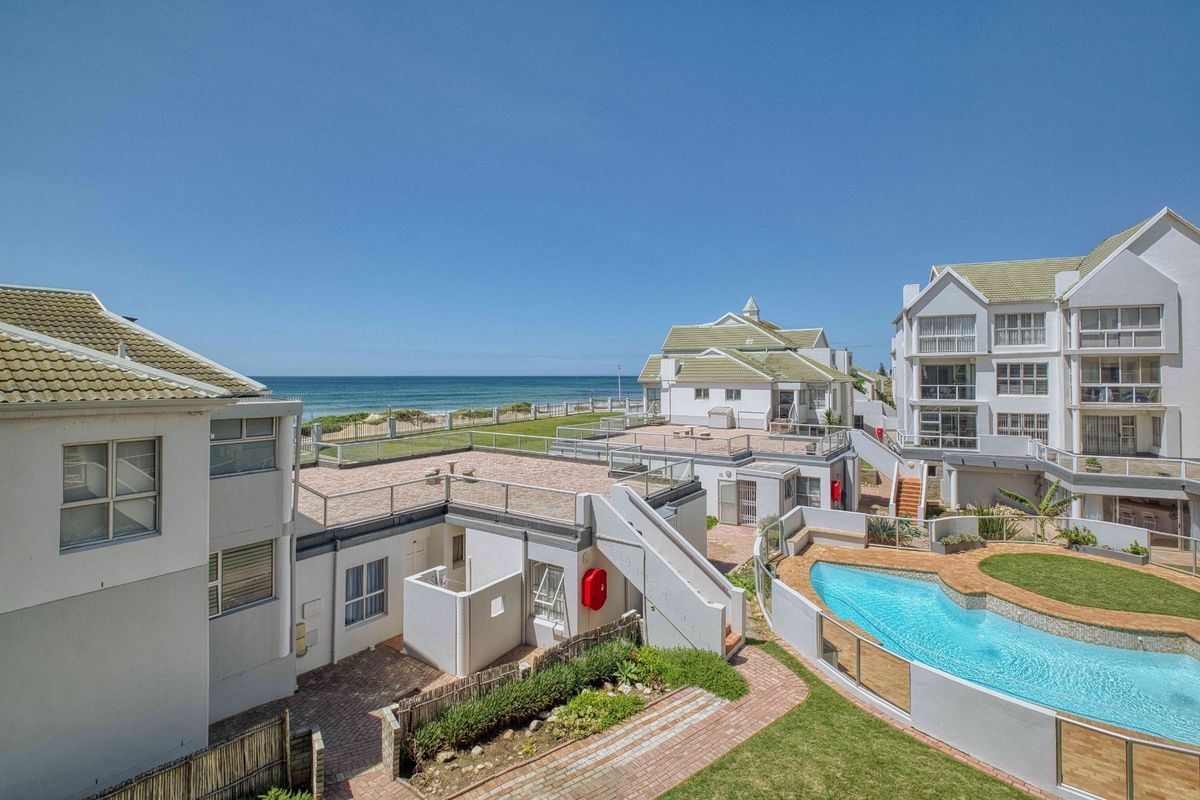 An elevated view of the apartment complex exterior, featuring a communal swimming pool, paved patio areas, and direct views of the ocean.