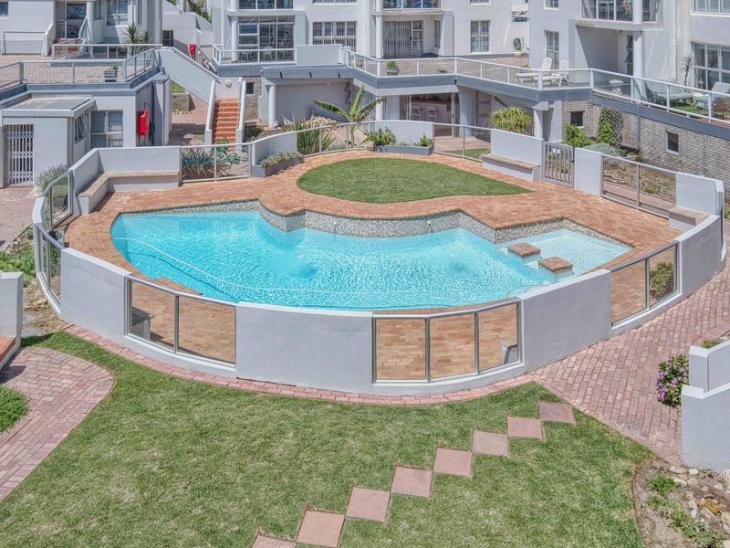 A well-maintained communal swimming pool area featuring a paved deck, glass safety fencing, and landscaped lawn within an apartment complex.