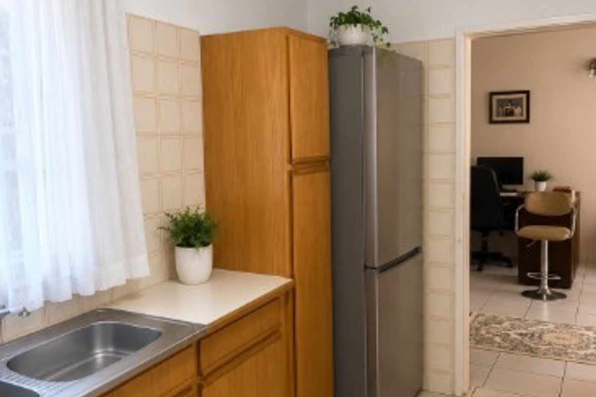 A functional kitchen featuring wooden cabinetry, a stainless steel double sink, and neutral ceramic tile flooring, with an adjacent view into a home office space.