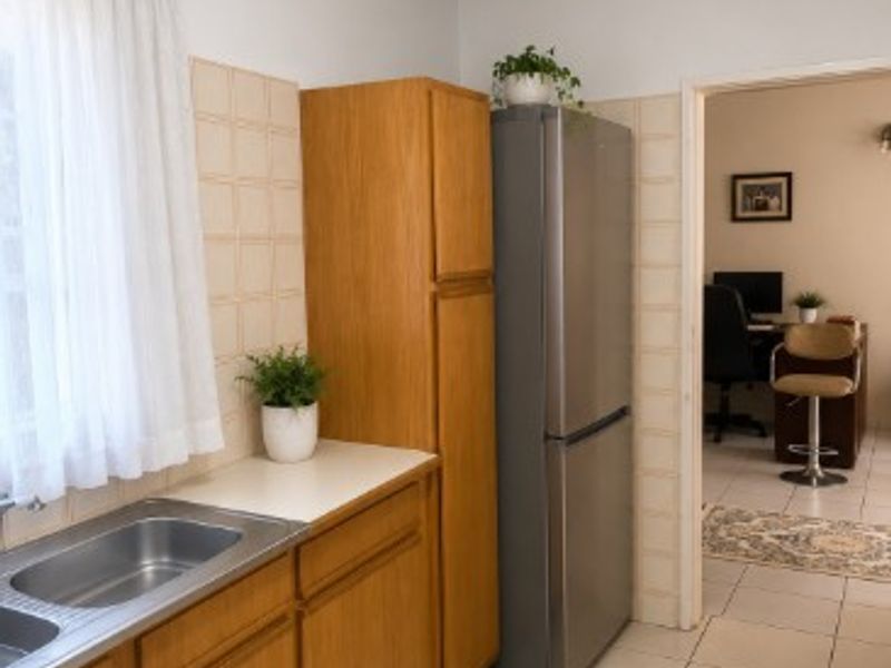 A functional kitchen featuring wooden cabinetry, a stainless steel double sink, and neutral ceramic tile flooring, with an adjacent view into a home office space.