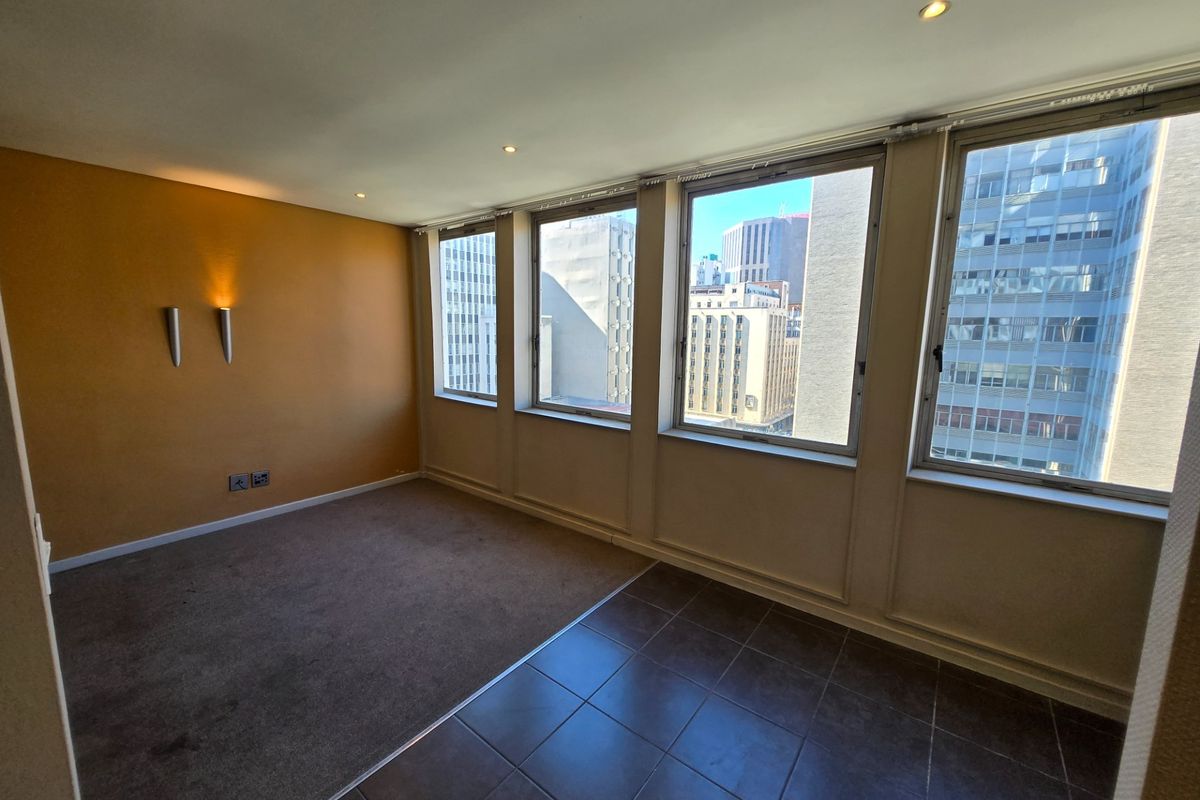 An empty living area featuring a mix of carpet and tiled flooring, recessed lighting, and large windows offering an urban city view.