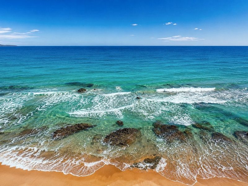 A scenic, elevated view of the ocean coastline featuring clear turquoise water, sandy beach, and rocky outcrops.