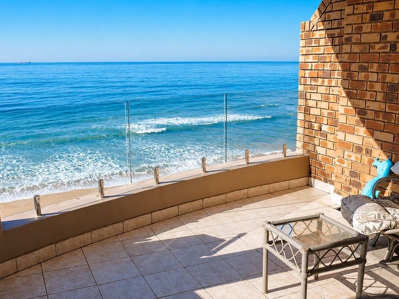 A spacious balcony featuring a modern glass balustrade, tiled flooring, and an exposed brick wall, offering direct, unobstructed views of the ocean.