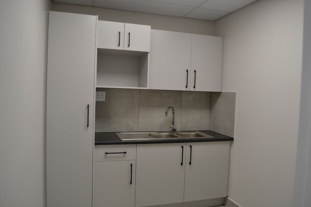 A clean, modern kitchenette featuring white built-in cabinetry, a stainless steel sink, and neutral tiled splashback, suitable for office or commercial use.