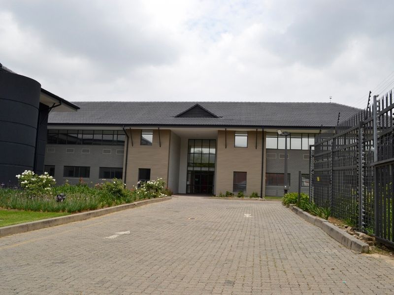 Front view of a commercial office building featuring a paved driveway, secure perimeter fencing, and a modern tiled roof facade.