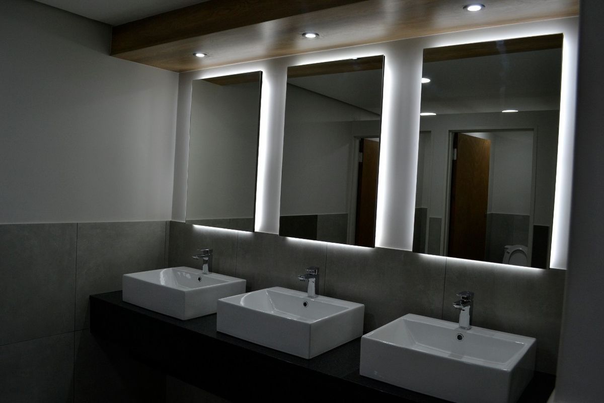 Modern commercial bathroom featuring a triple vanity with vessel sinks, sleek backlit mirrors, and contemporary tiled finishes.