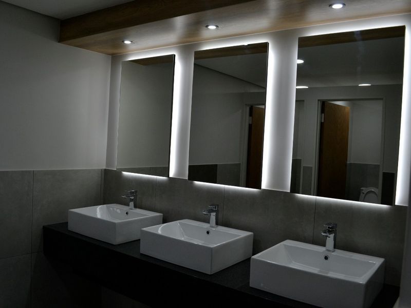 Modern commercial bathroom featuring a triple vanity with vessel sinks, sleek backlit mirrors, and contemporary tiled finishes.
