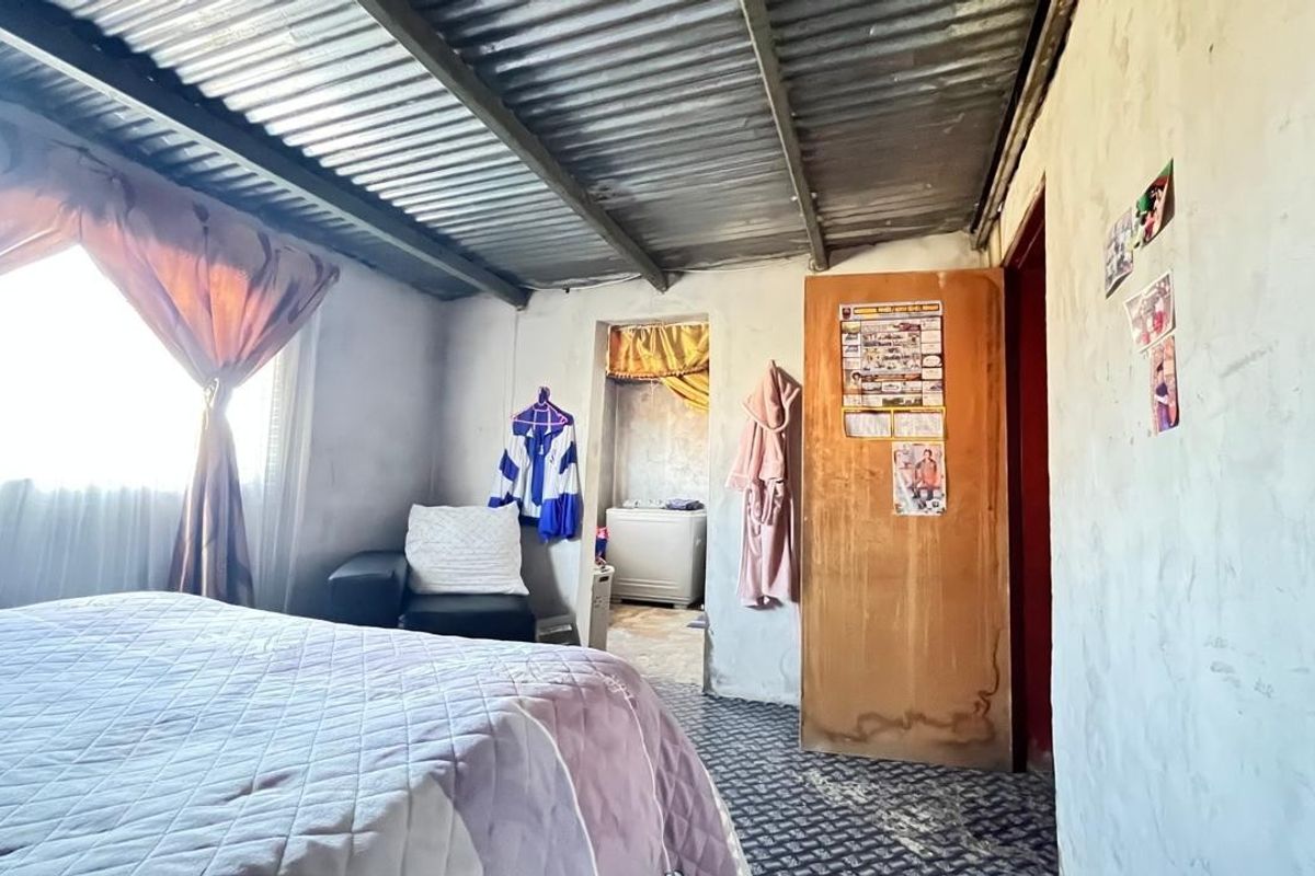 A bedroom featuring a corrugated metal ceiling, patterned vinyl flooring, and a simple timber door, offering a basic living space.