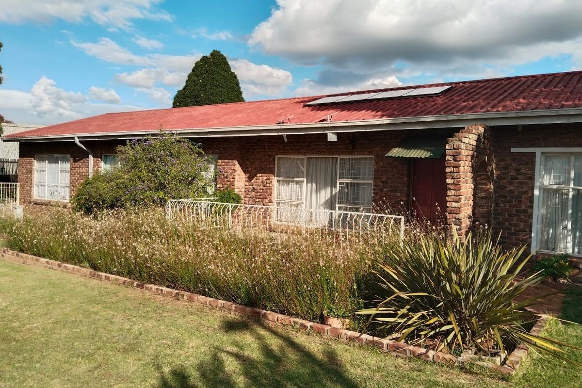 A single-story face brick house featuring a red corrugated metal roof with solar panels and a paved patio area with security fencing.