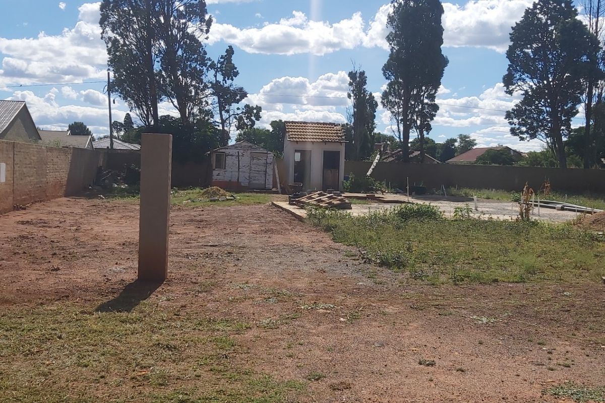 A vacant residential plot featuring a perimeter brick wall, a concrete foundation slab, and an existing outbuilding structure.