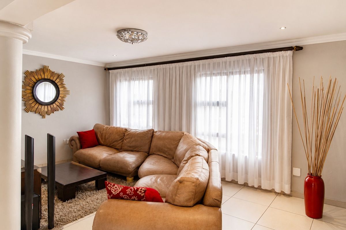 A well-maintained living room featuring neutral tiled flooring, recessed lighting, and large windows dressed with sheer curtains.