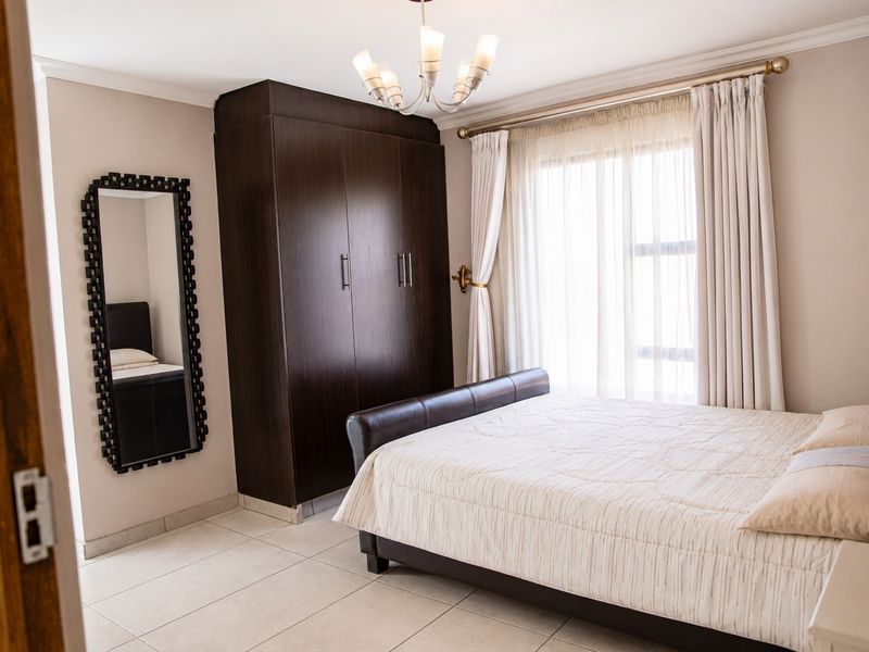 A well-maintained bedroom featuring neutral tiled flooring, a dark wood built-in wardrobe, and natural light through large windows with elegant drapery.
