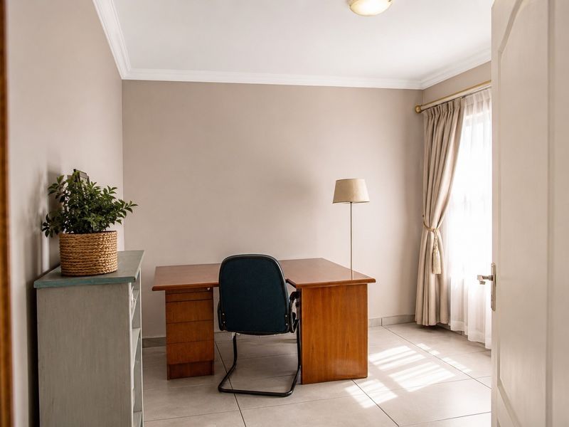 A bright home office featuring neutral wall tones, tiled flooring, and natural light through a window with fitted curtains.