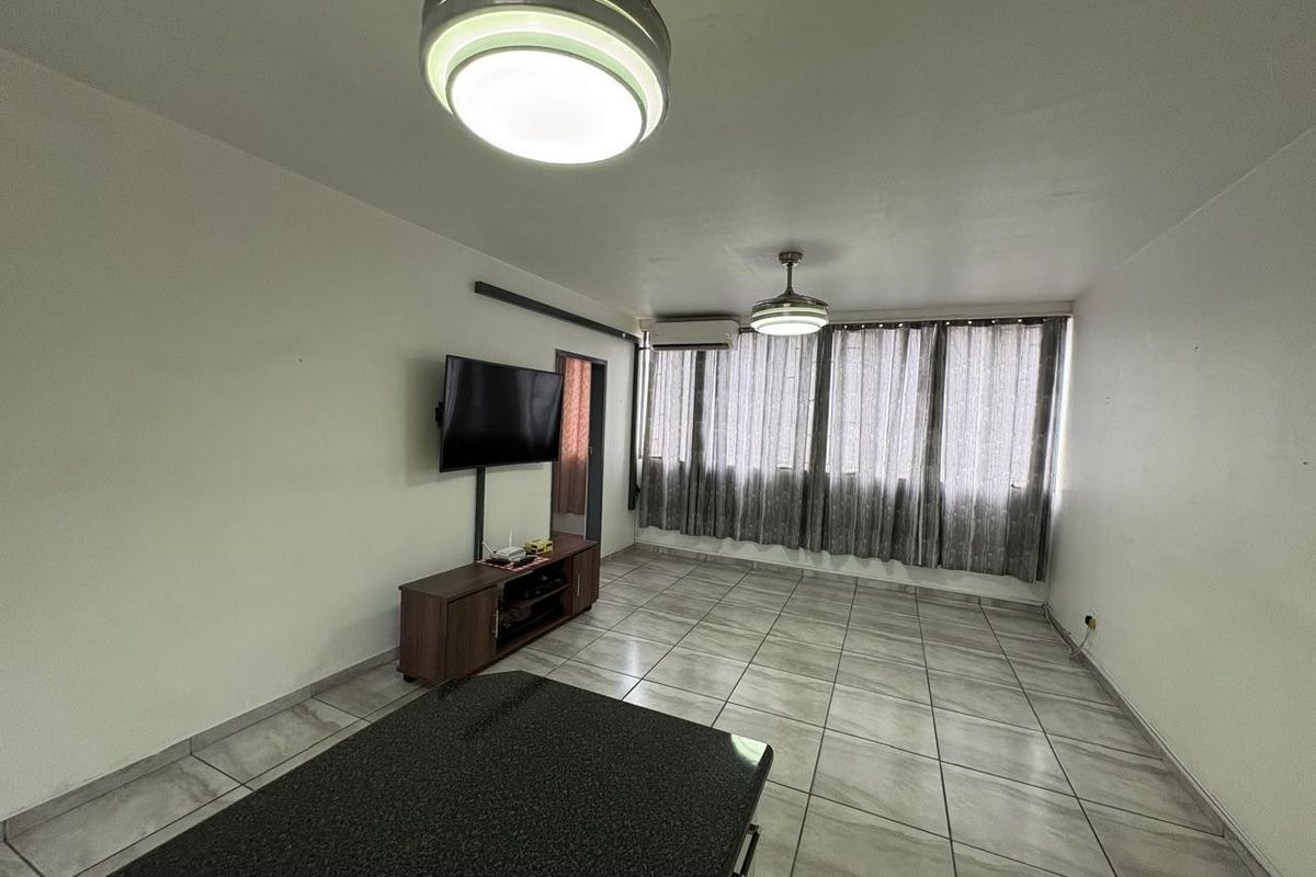 A living room featuring tiled flooring, a ceiling fan, and a wall-mounted air conditioning unit, offering a functional and neutral space.