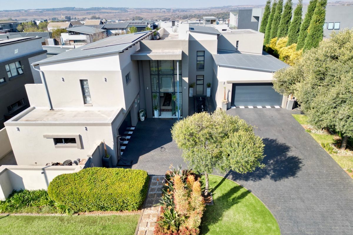 Aerial view of a modern luxury residence featuring a paved driveway, double garage, solar panels, and a striking glass-fronted entrance.