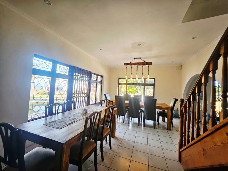 Spacious dining area featuring ceramic floor tiles, recessed lighting, and a wooden staircase, with large windows providing natural light and garden views.
