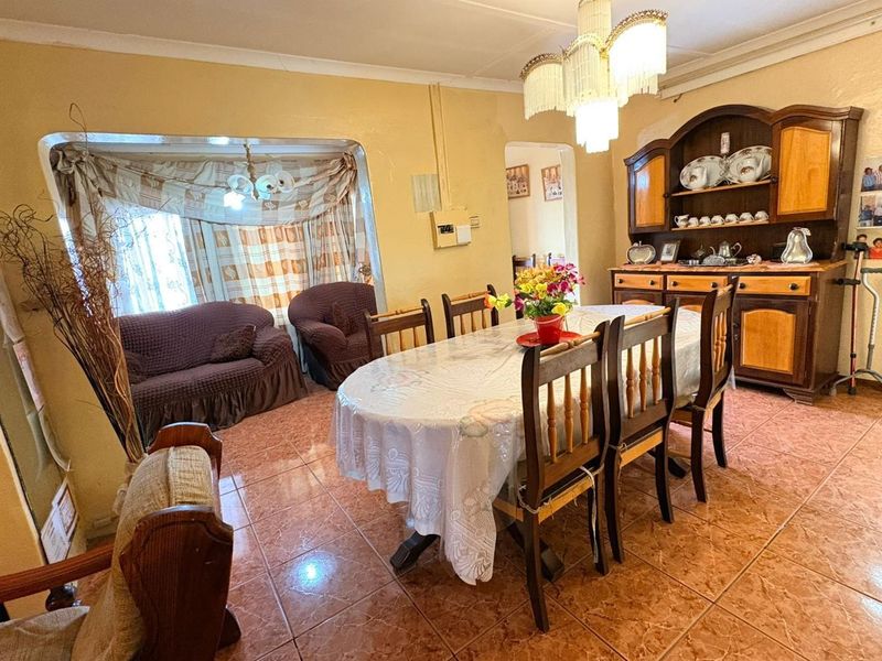 A dining room featuring tiled flooring and a central chandelier, with an adjacent living area visible through an arched opening.