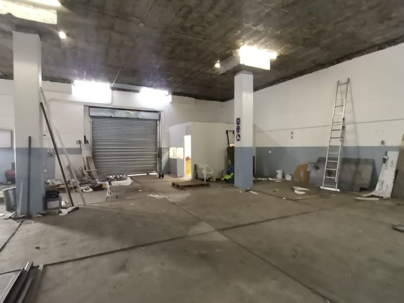 Spacious industrial utility area featuring a high concrete ceiling, concrete flooring, structural columns, and a large roller shutter door for easy access.