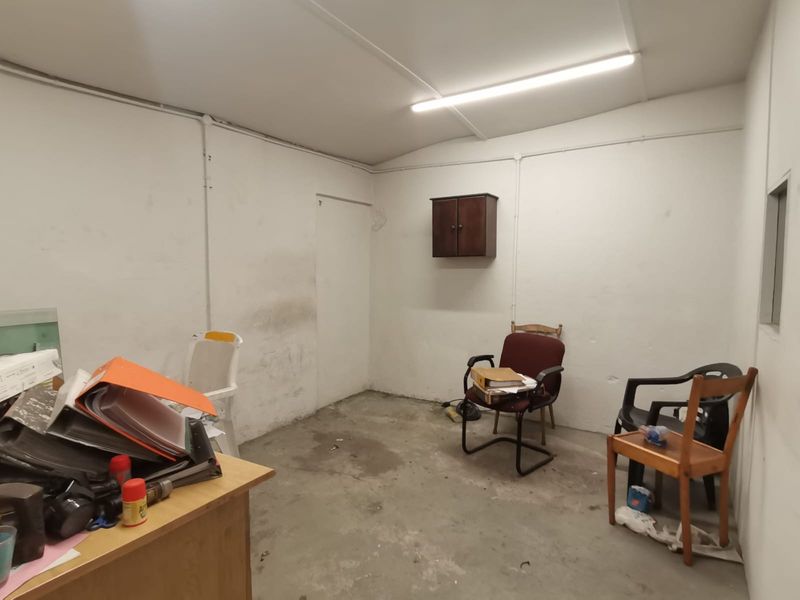 A basic industrial office space featuring exposed electrical conduit, fluorescent lighting, and a wall-mounted cabinet, requiring cosmetic updates to the walls and flooring.