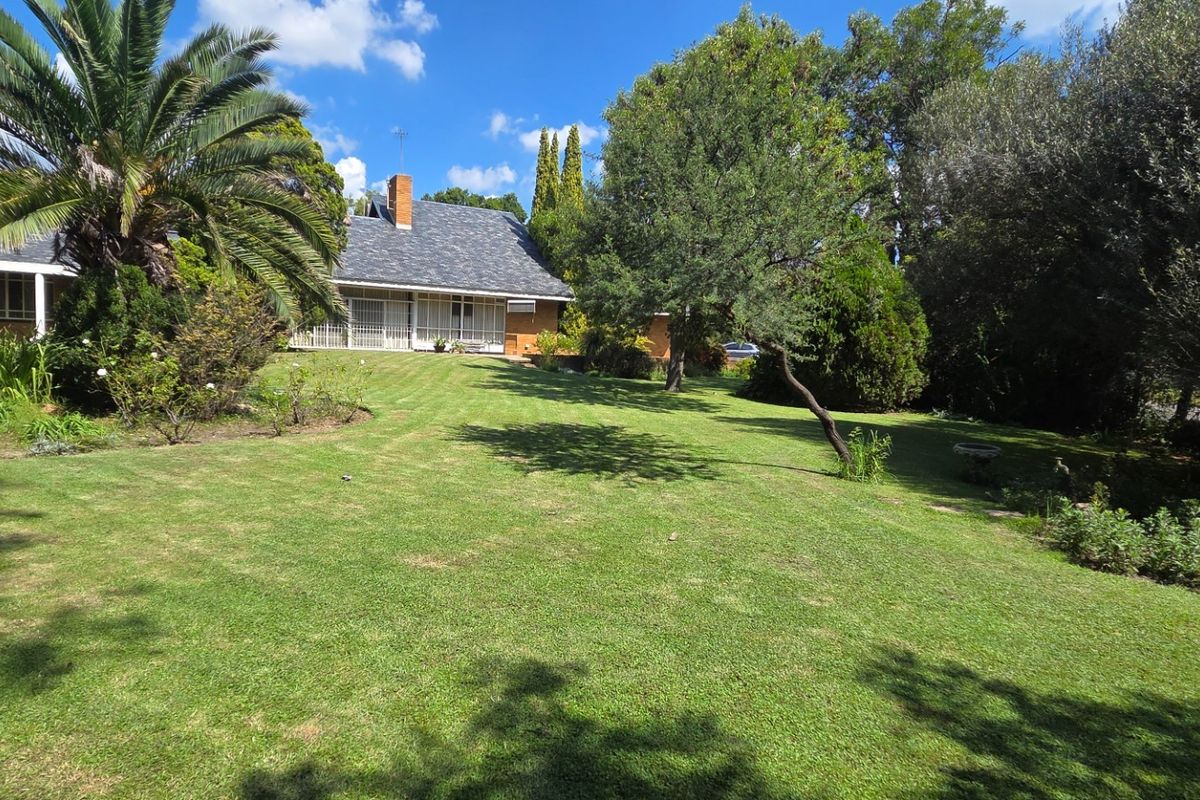 A spacious rear garden featuring a well-maintained lawn, mature trees, and a view of the house with a brick chimney and covered patio area.