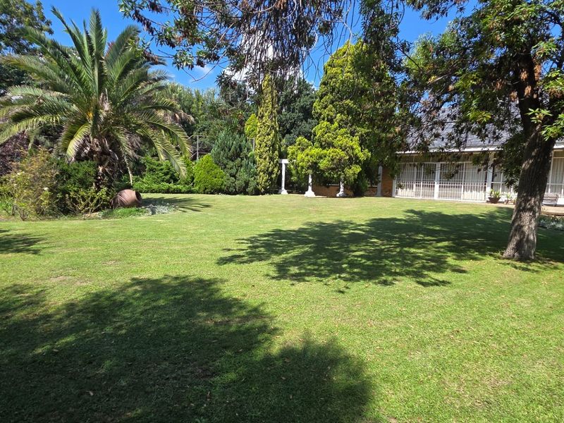 A spacious, well-maintained garden featuring a large lawn, mature trees, and a palm tree, with a covered patio area visible in the background.