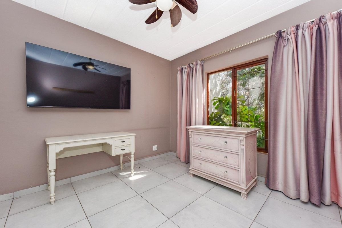 A bedroom featuring neutral wall tones, tiled flooring, and a ceiling fan, with a window providing natural light and garden views.