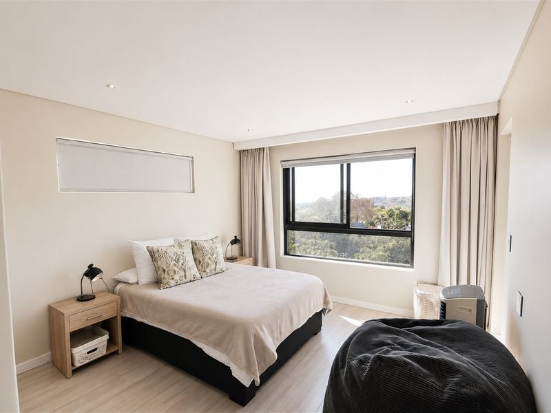 A bright and modern bedroom featuring neutral tones, laminate flooring, recessed lighting, and a large window with views of the surrounding greenery.