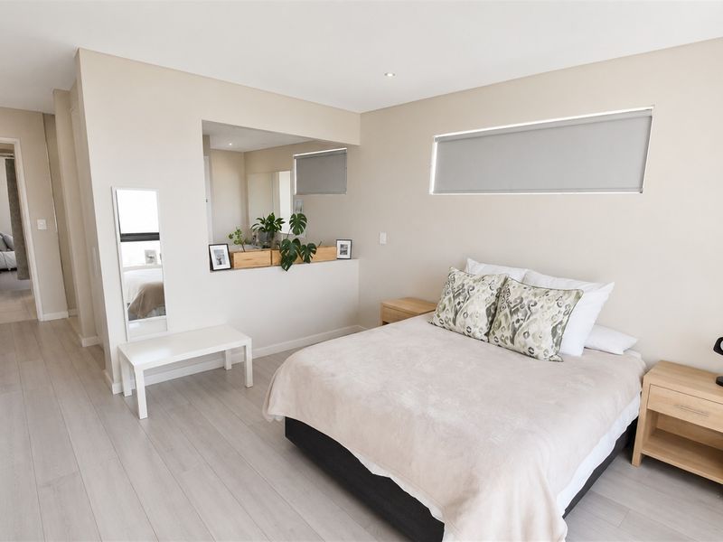 A bright and modern bedroom featuring neutral tones, laminate flooring, recessed lighting, and an internal window providing a view into the adjacent space.