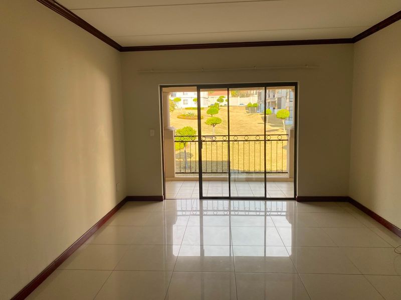 An empty, well-maintained living room featuring neutral tiled floors, crown molding, and a sliding glass door leading to a private balcony with garden views.