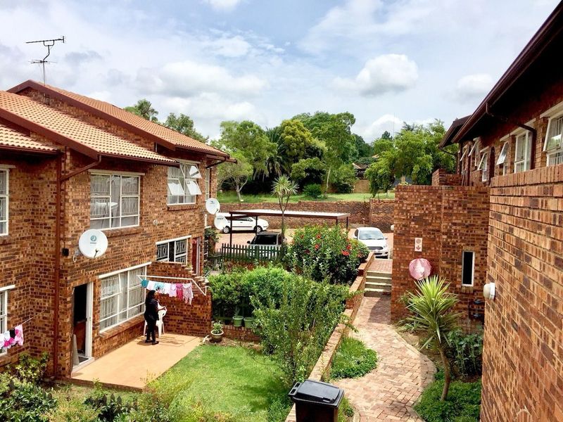 A view of the rear exterior of the apartment complex, featuring face brick buildings, a paved walkway, landscaped garden areas, and a communal carport.